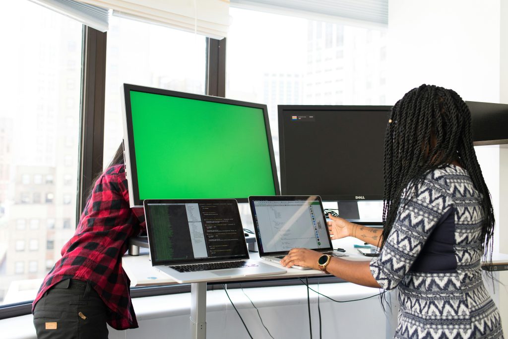 Two women working on laptops and monitors in a bright office setting, focused on technology and teamwork.